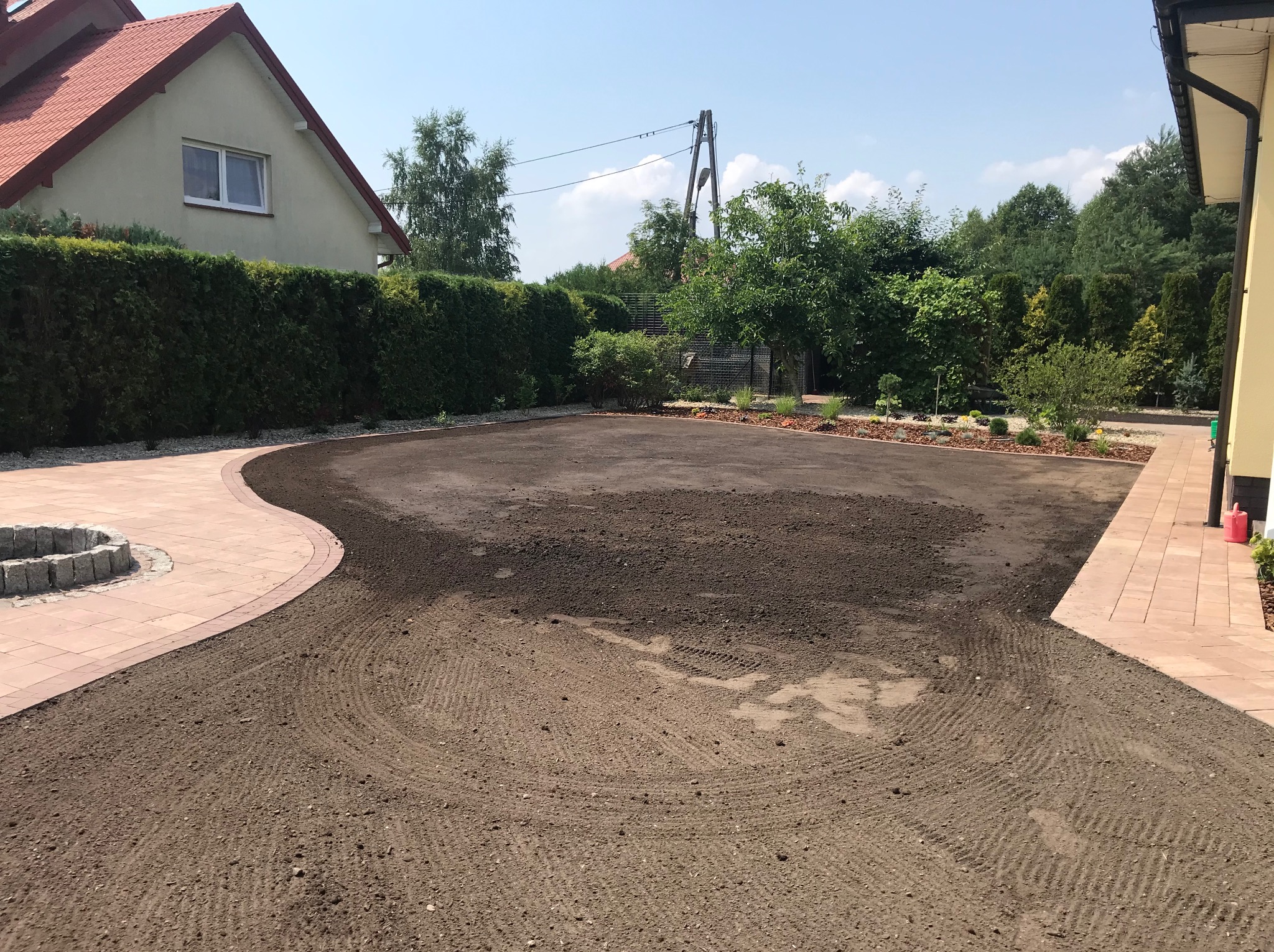 Przygotowany teren pod trawnik z widocznymi śladami po bronowaniu, obrzeża z kostki brukowej i fragment domu w tle, słoneczny dzień.