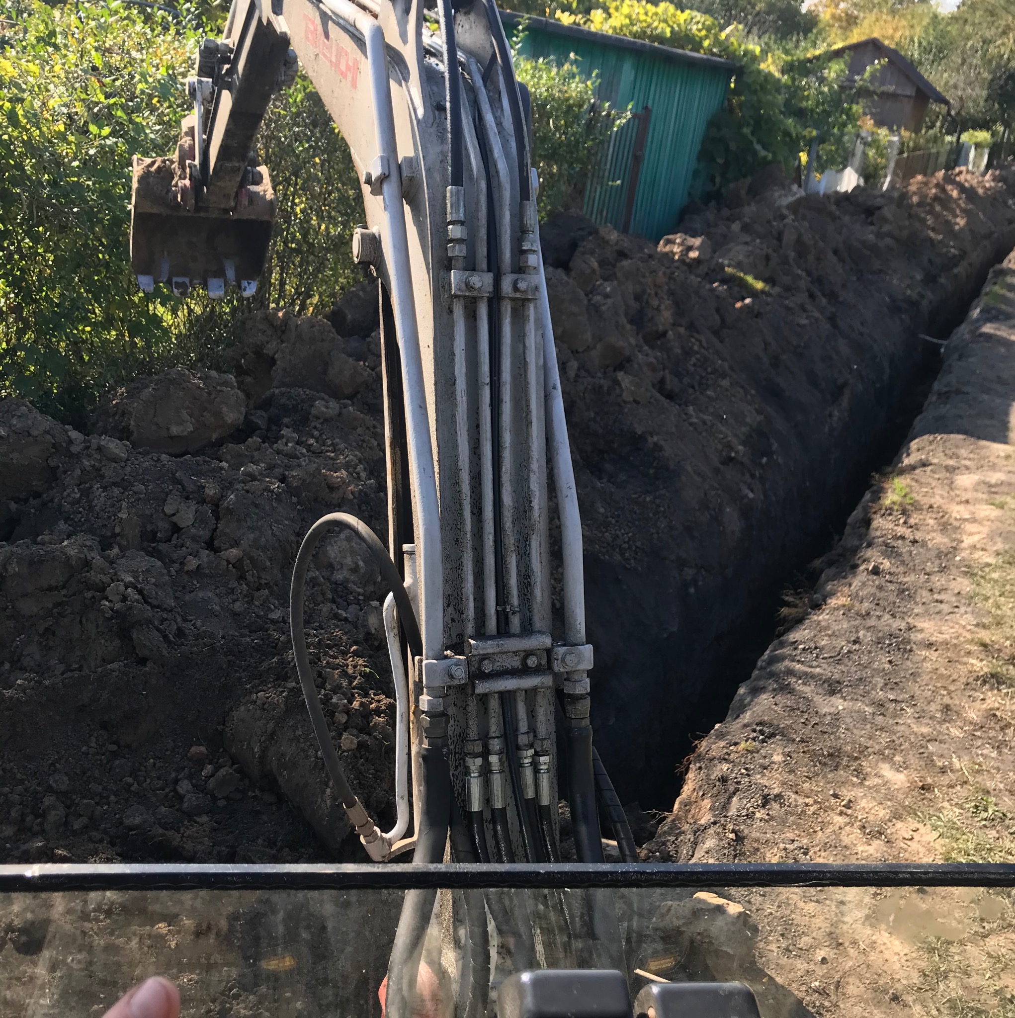 Koparka wykonuje wykop liniowy w ziemi, widok z kabiny operatora, zarośla w tle. Wykop pod instalacje lub fundamenty.