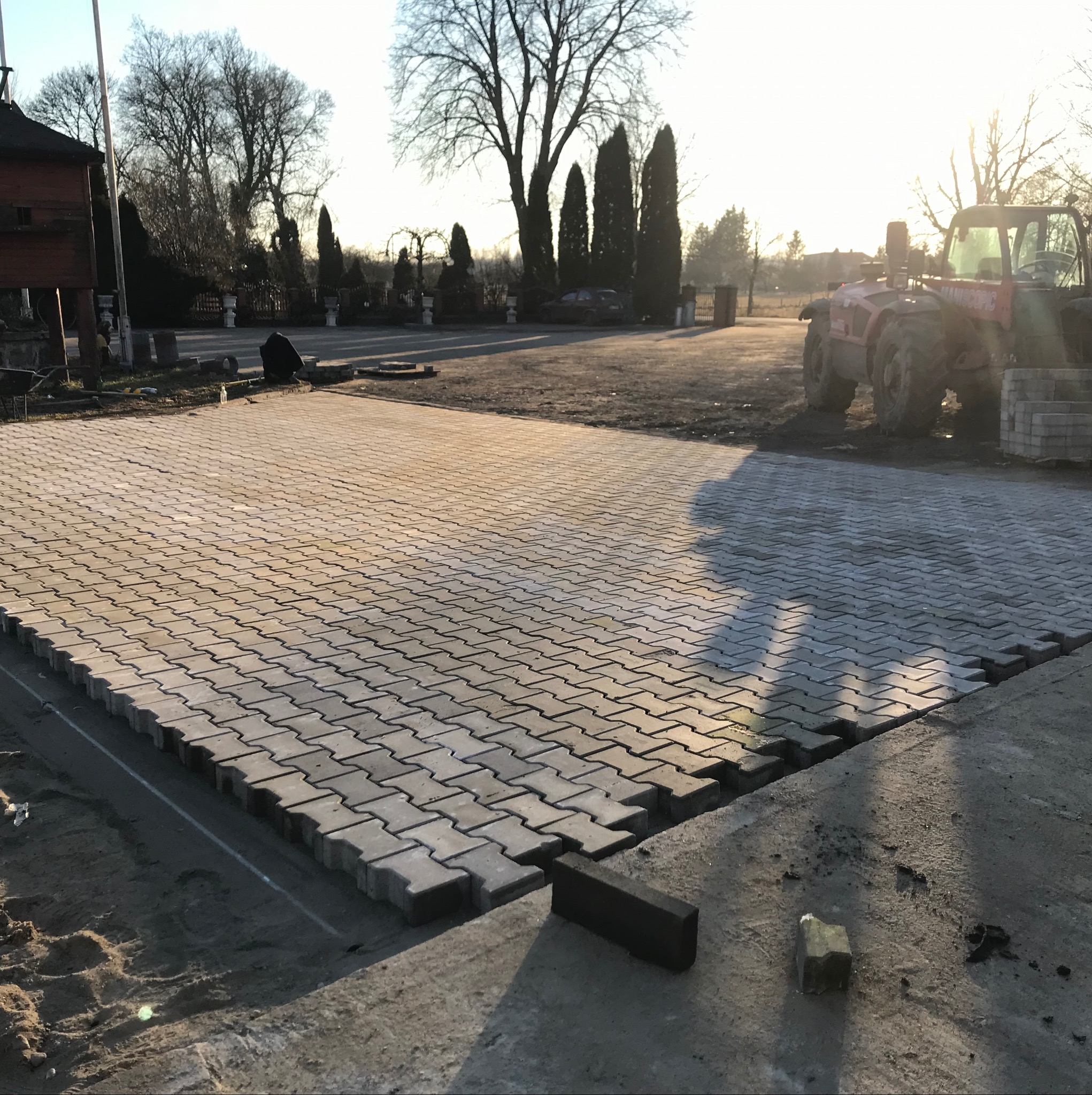 Trwa układanie szarej kostki brukowej w Łyse, widoczny fragment placu i maszyna budowlana w tle. Praca w toku, widoczne niedokończone krawędzie i pojedyncze kostki.