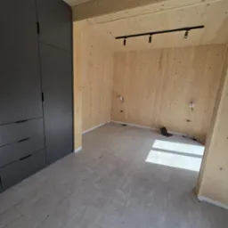 Wykończone wnętrze z drewnianymi ścianami, szarą zabudową meblową i panelami podłogowymi. Widoczne oświetlenie sufitowe i przygotowane punkty elektryczne.