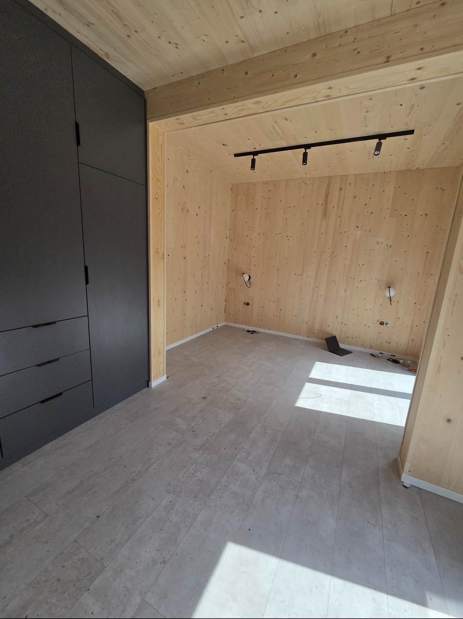 Wykończone wnętrze z drewnianymi ścianami, szarą zabudową meblową i panelami podłogowymi. Widoczne oświetlenie sufitowe i przygotowane punkty elektryczne.