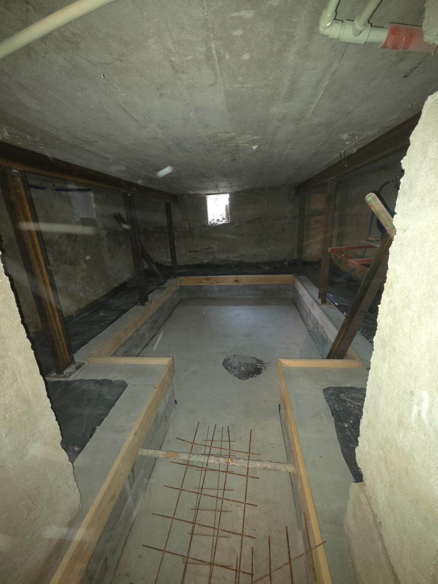 Przygotowanie betonowej wylewki w piwnicy: szalunek z desek, zbrojenie z drutu, izolacja. Widoczne stalowe słupy wzmacniające konstrukcję.