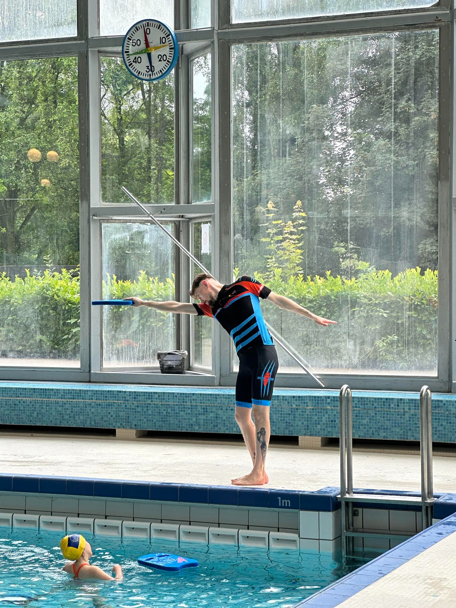 Instruktor pływania demonstruje ćwiczenie z deską na brzegu basenu, w tle zegar i duże okno z widokiem na zieleń, w wodzie pływa dziecko w czepku.