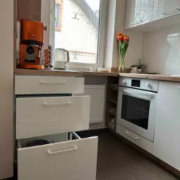 Nowoczesna, jasna kuchnia z białymi, lakierowanymi frontami, drewnianym blatem, widocznym piekarnikiem i częściowo wysuniętą szufladą, na blacie stoi pomarańczowy ekspres do kawy i wazon z tulipanami.