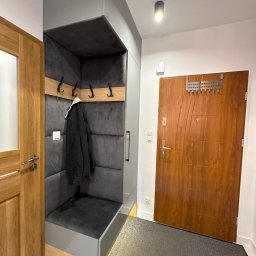 Mateusz Stano - Nowoczesny przedpokój z zabudową meblową: szara szafka z siedziskiem i wieszakami, drewniane drzwi wewnętrzne i wejściowe, szary dywanik przy drzwiach wejściowych.