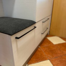 Mateusz Stano - Fragment szafy wnękowej z siedziskiem i szufladami, jasne drewno, czarne uchwyty, na podłodze brązowe płytki.