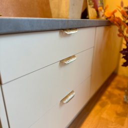 Mateusz Stano - Białe, gładkie fronty szuflad z dekoracyjnymi, złotymi uchwytami w kształcie listków na tle drewnianej podłogi i blatu z szarego kamienia.