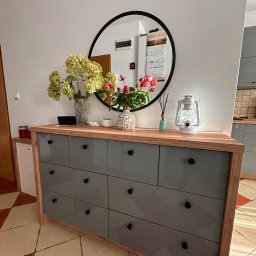 Mateusz Stano - Szara komoda z drewnianym blatem i czarnymi uchwytami, na blacie wazony z kwiatami i lampion dekoracyjny, okrągłe lustro w tle, płytki podłogowe w romby.