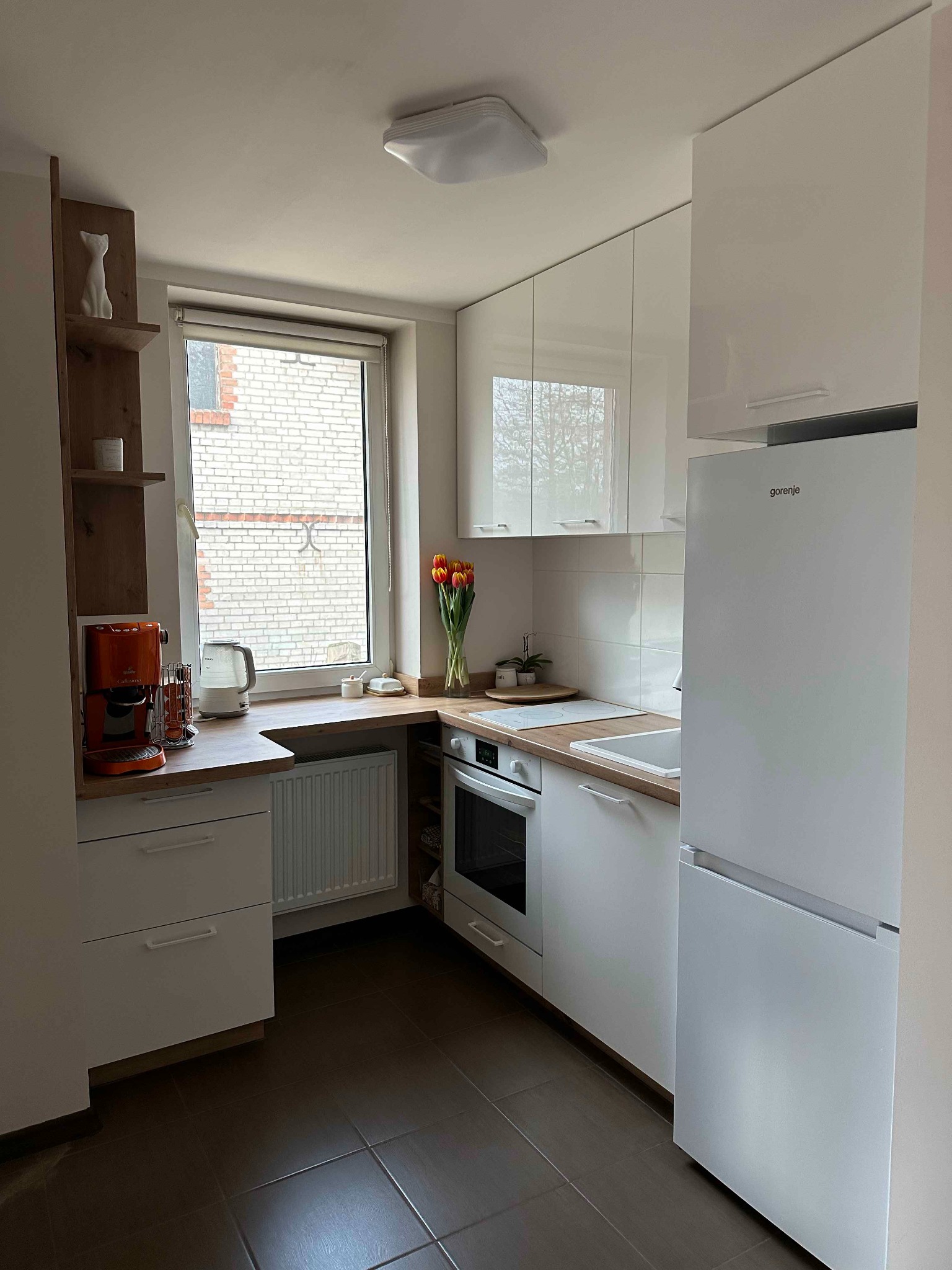 Jasna kuchnia z białymi lakierowanymi frontami, brązowymi płytkami na podłodze i drewnianym blatem, pomarańczowy ekspres do kawy na tle okna.