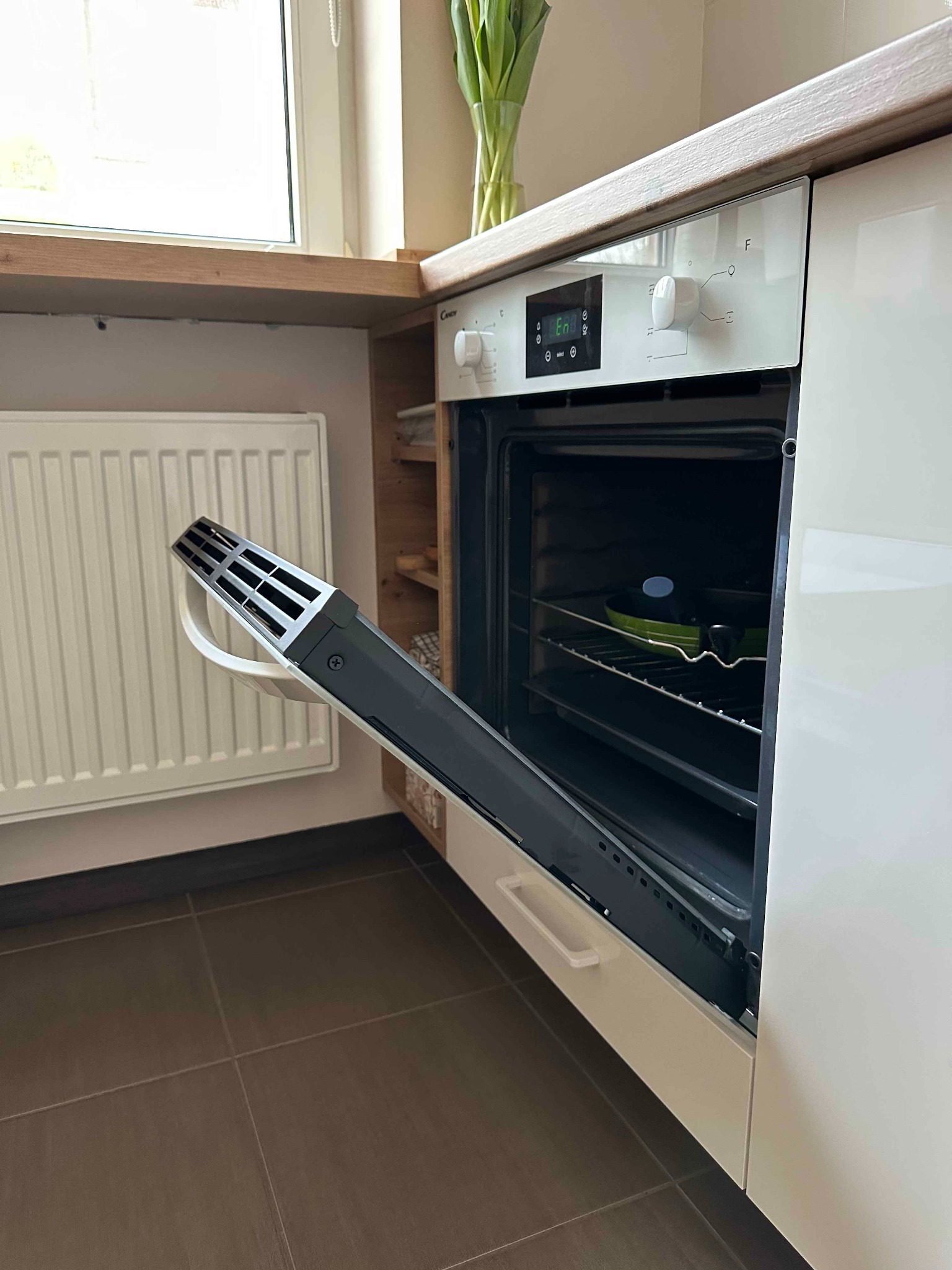 Otwarty piekarnik marki Candy w zabudowie kuchennej z białymi frontami, widoczna zielona patelnia w środku, obok grzejnik i brązowe płytki podłogowe.