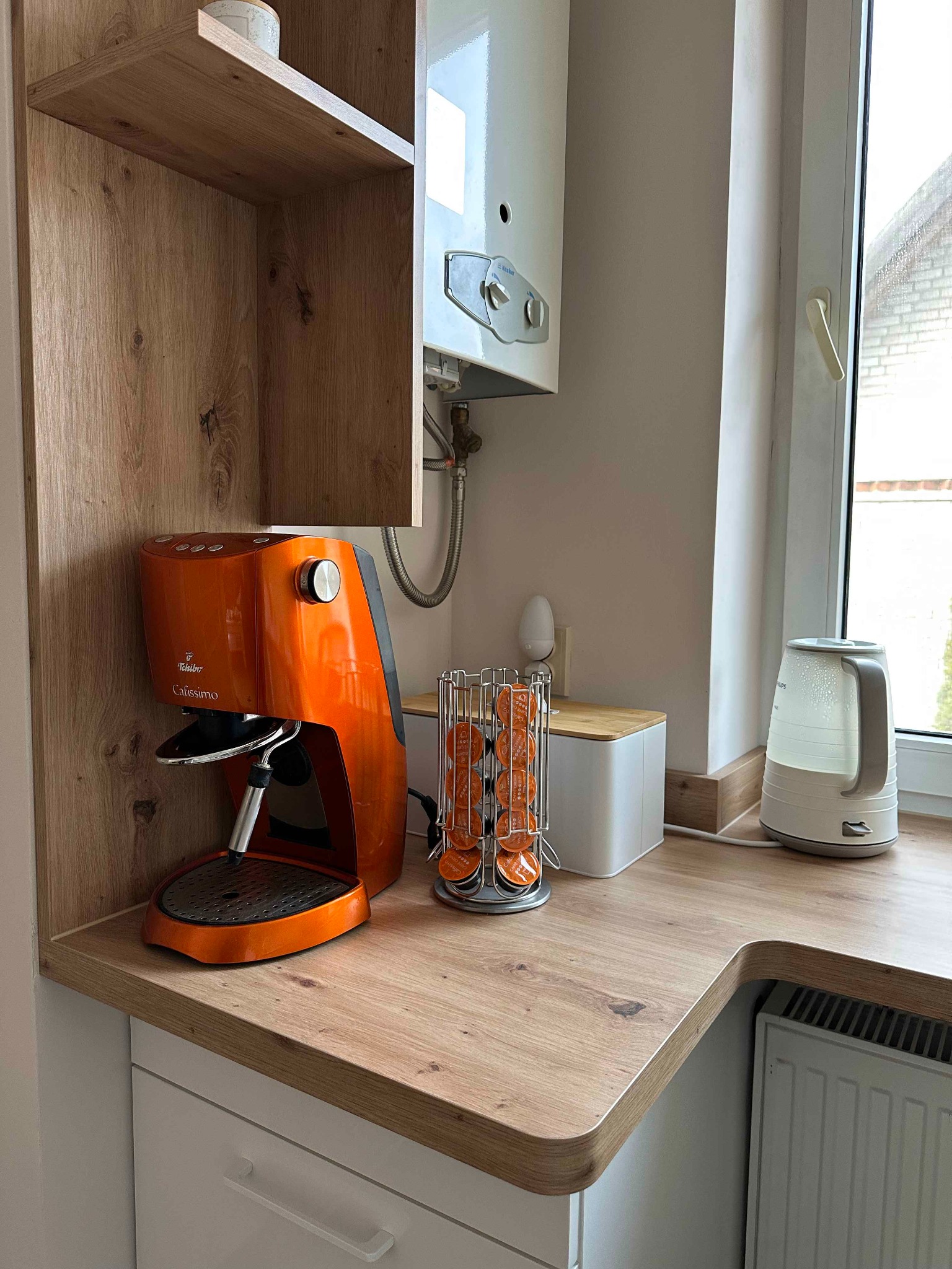 Fragment kuchni z drewnianym blatem, na którym stoi pomarańczowy ekspres do kawy Tchibo Cafissimo, obok metalowy stojak z kapsułkami, biały czajnik elektryczny oraz wiszący na ścianie biały piec...
