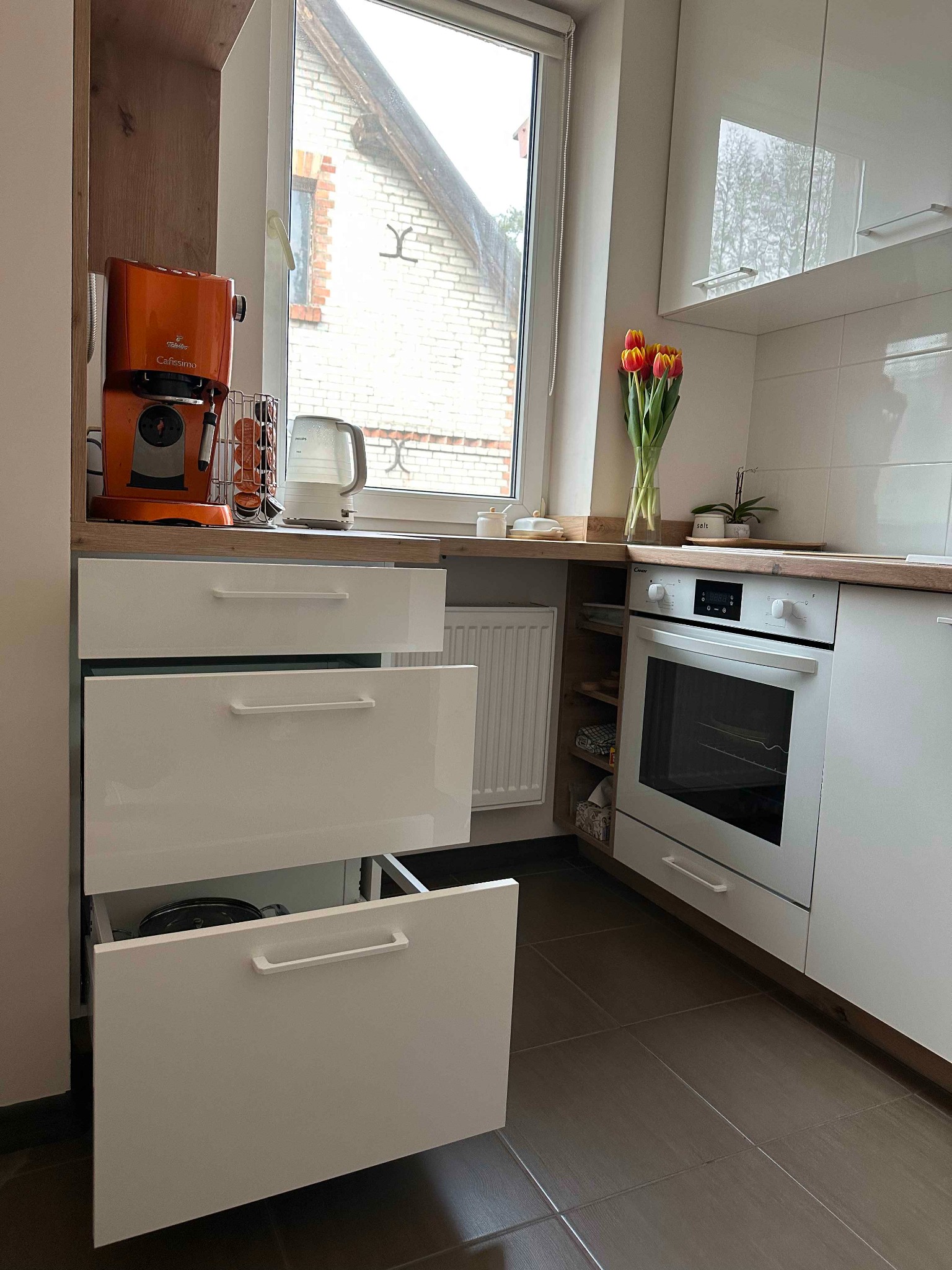 Nowoczesna, jasna kuchnia z białymi, lakierowanymi frontami, drewnianym blatem, widocznym piekarnikiem i częściowo wysuniętą szufladą, na blacie stoi pomarańczowy ekspres do kawy i wazon z tulipanami.