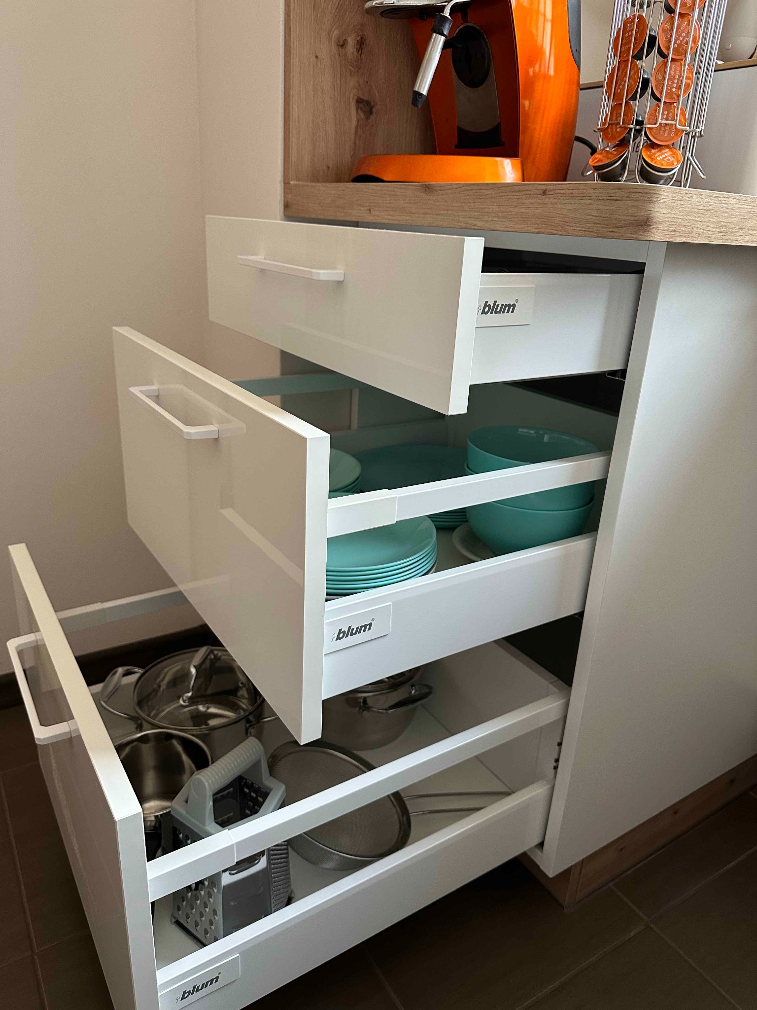 Otwarta szafka kuchenna z trzema wysuwanymi szufladami marki Blum, wypełnionymi naczyniami i akcesoriami kuchennymi, w tle pomarańczowy ekspres do kawy.
