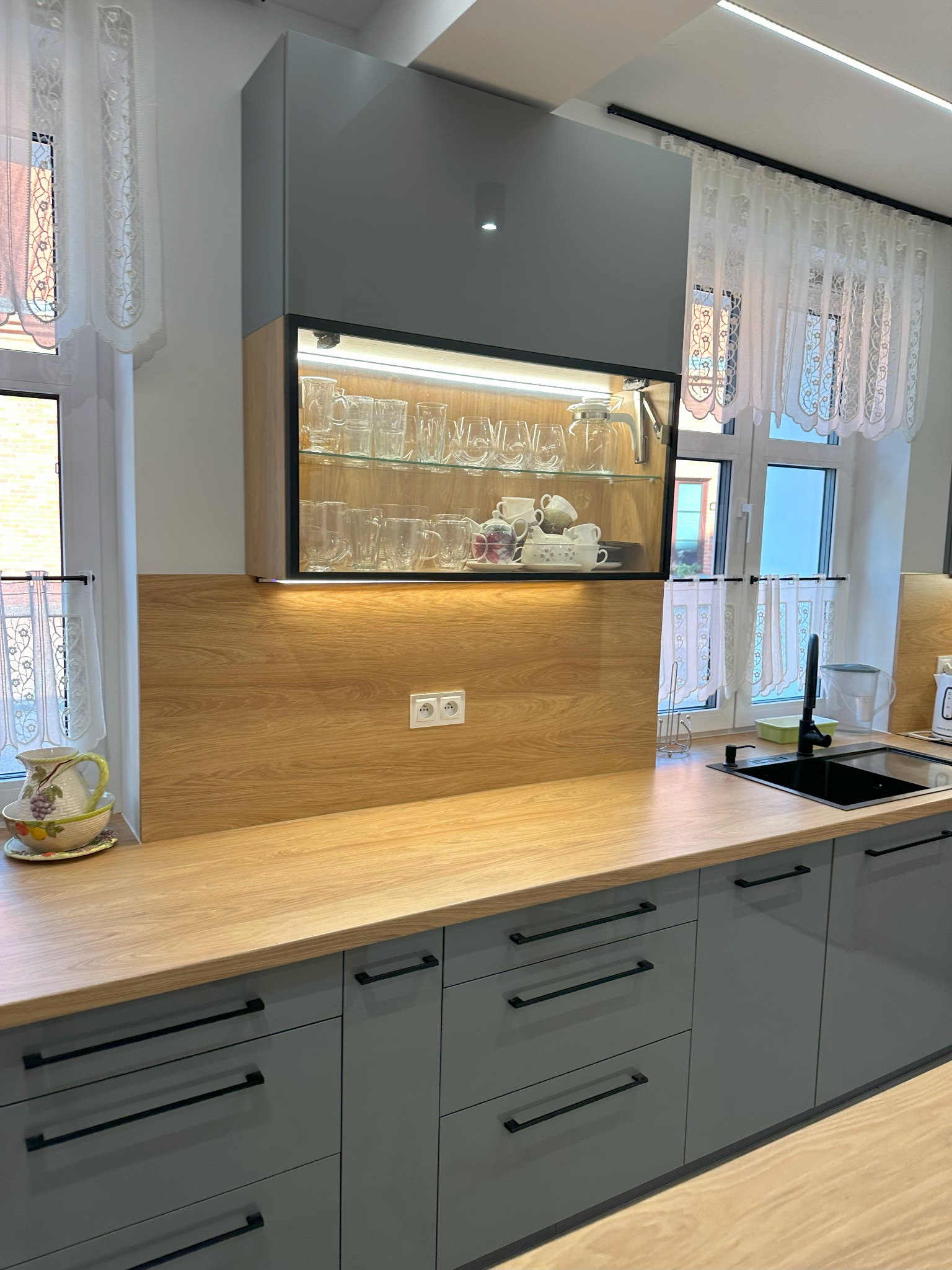 Szare szafki kuchenne z drewnianym blatem i przeszkloną szafką wiszącą z oświetleniem LED, zastawa stołowa w szafce, ozdobny dzbanek na blacie, okno z firanką w tle.