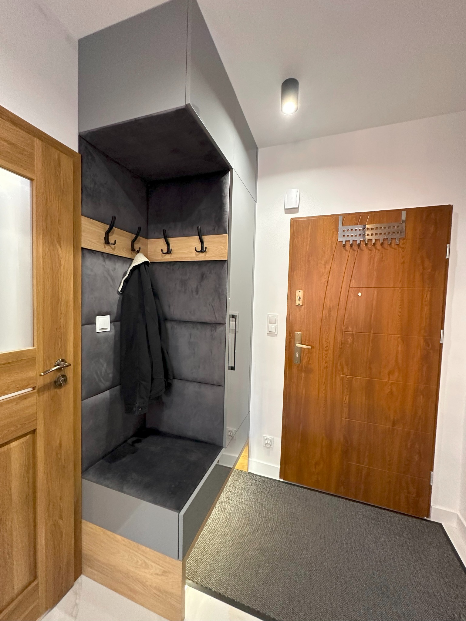 Nowoczesny przedpokój z zabudową meblową: szara szafka z siedziskiem i wieszakami, drewniane drzwi wewnętrzne i wejściowe, szary dywanik przy drzwiach wejściowych.
