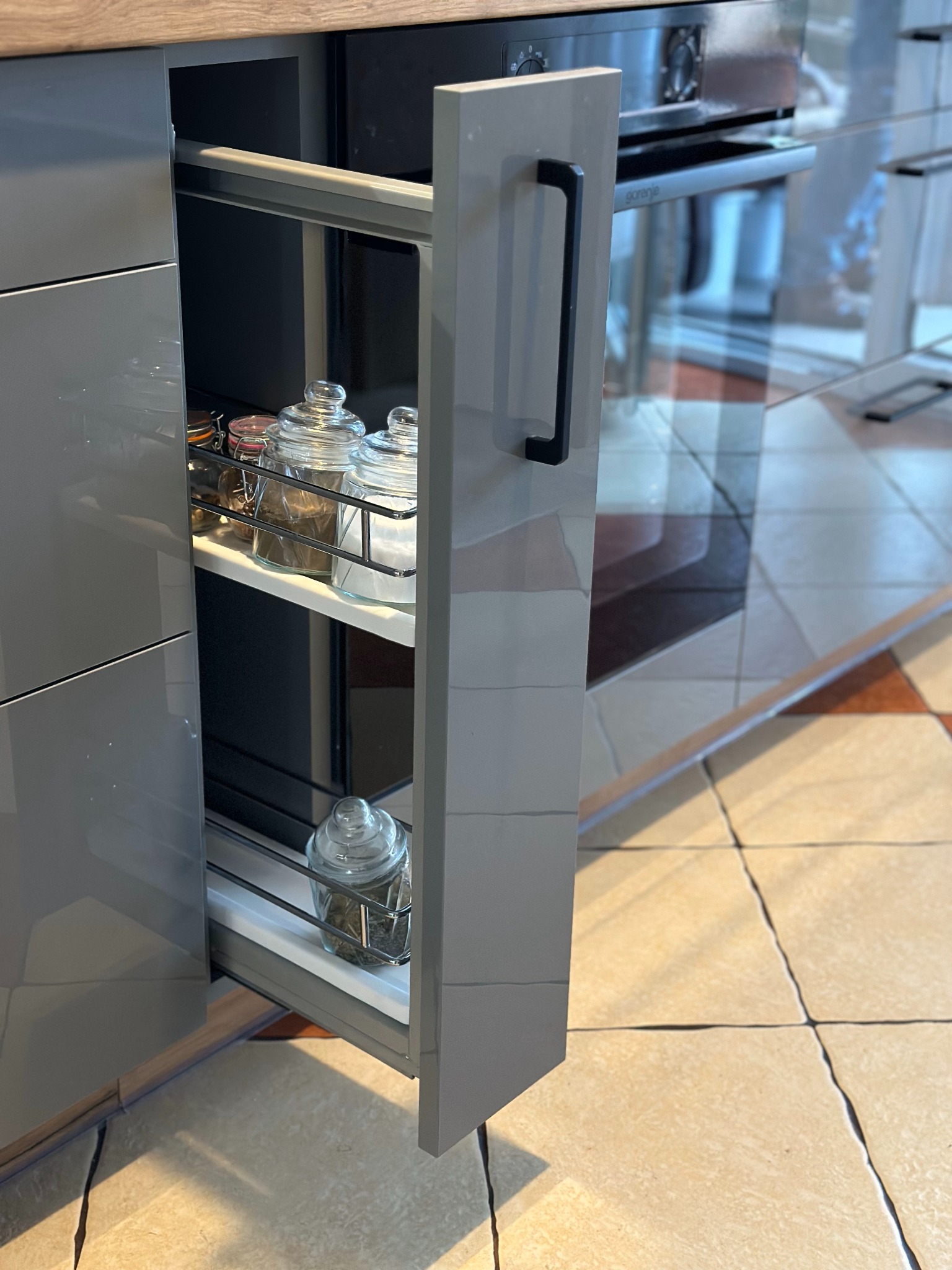 Wysuwana, szara szafka kuchenna z metalowymi relingami, wypełniona szklanymi pojemnikami na przyprawy, na tle płytek podłogowych i zabudowanego piekarnika.