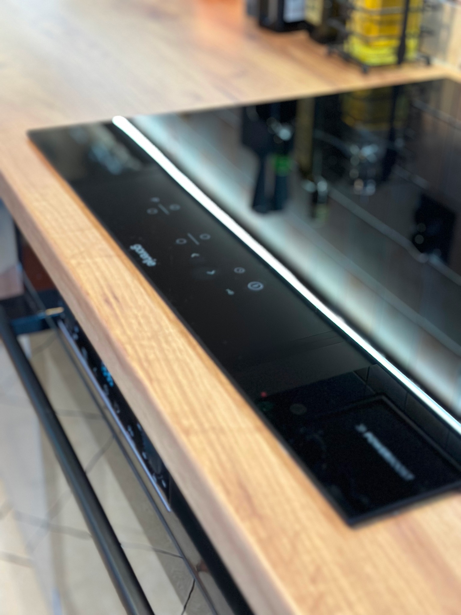 Czarna płyta indukcyjna Gorenje wbudowana w drewniany blat kuchenny, widoczne dotykowe panele sterowania i odbicie otoczenia.