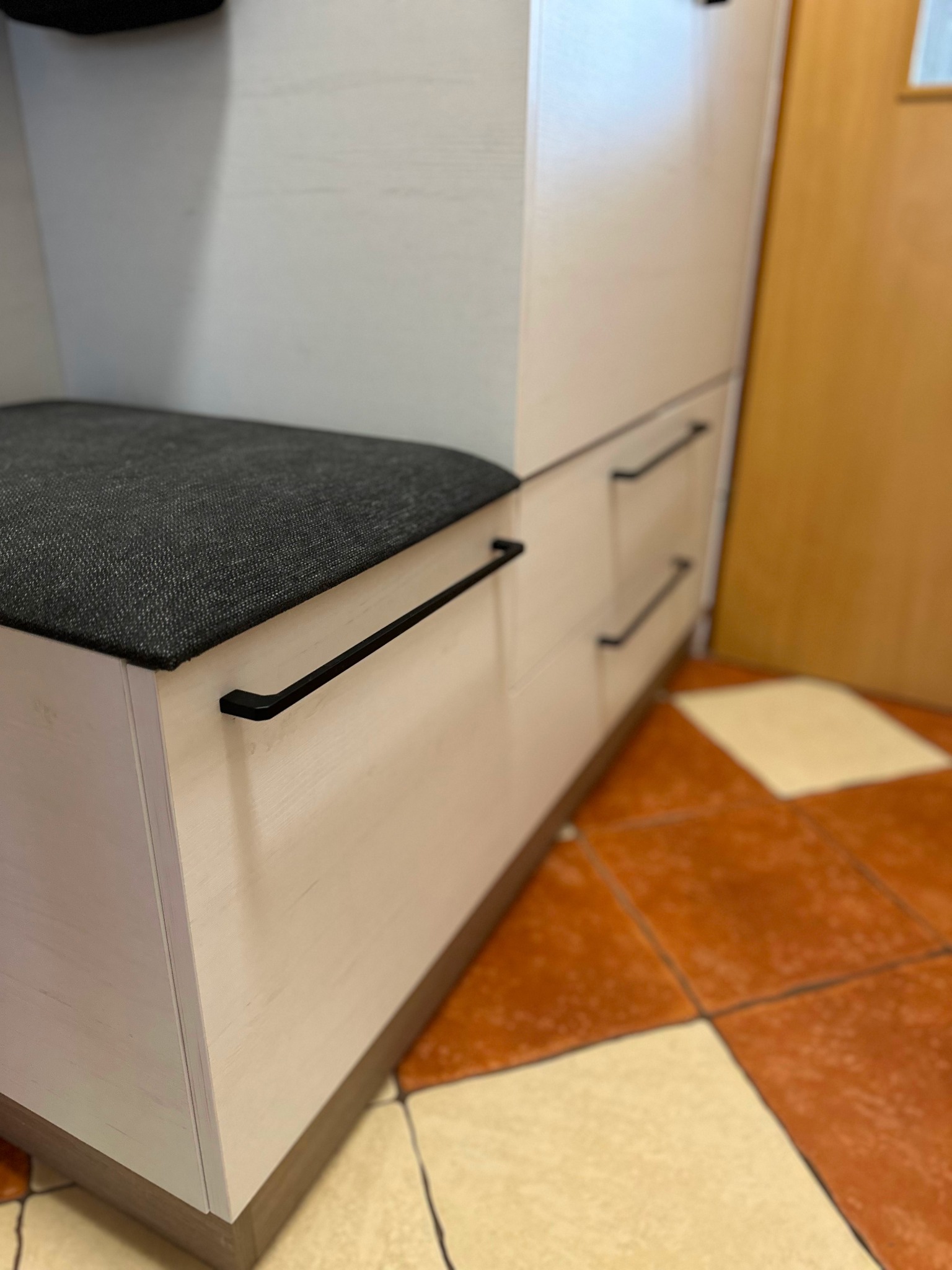 Fragment szafy wnękowej z siedziskiem i szufladami, jasne drewno, czarne uchwyty, na podłodze brązowe płytki.