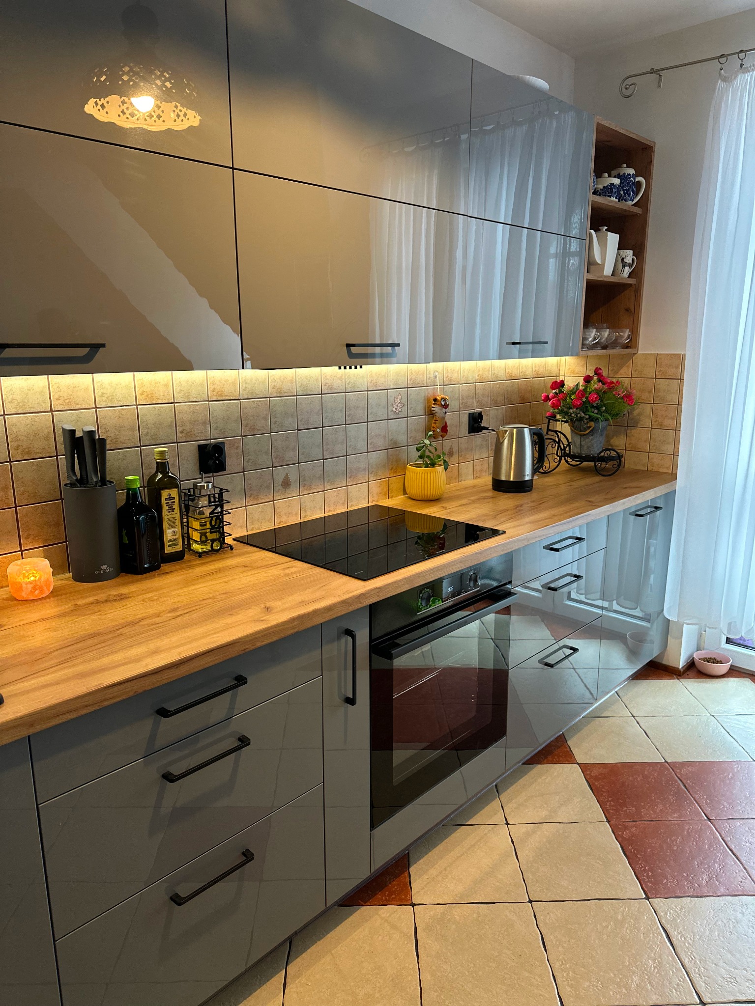 Szare, lakierowane szafki kuchenne z drewnianym blatem, podświetleniem LED i płytkami w tle; widoczny piekarnik, płyta indukcyjna, czajnik elektryczny i dekoracje.