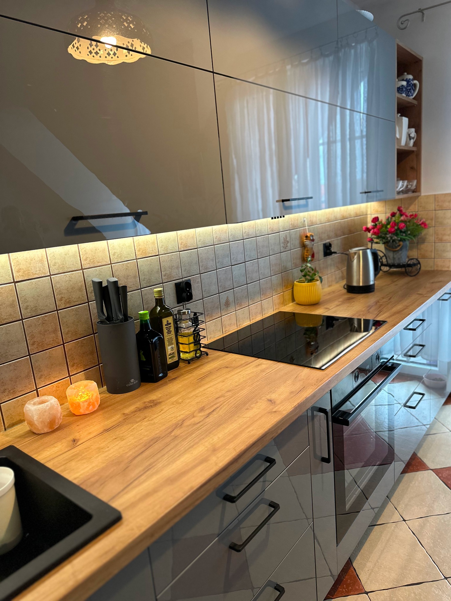 Nowoczesna kuchnia z szarymi, lakierowanymi frontami, drewnianym blatem, indukcyjną płytą grzewczą, oświetleniem podszafkowym LED i dekoracyjnymi akcentami, takimi jak himalajskie lampki solne.