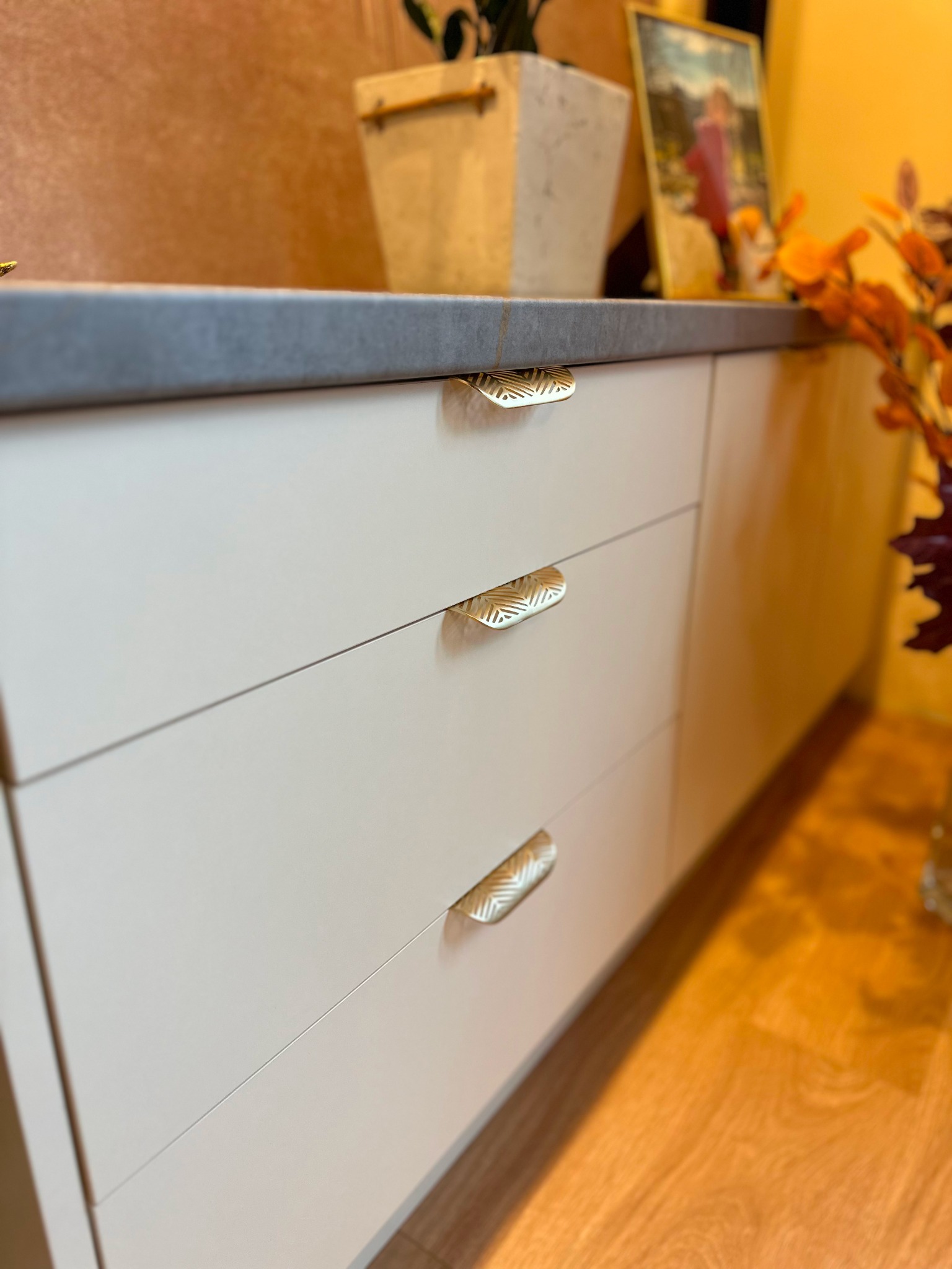 Białe, gładkie fronty szuflad z dekoracyjnymi, złotymi uchwytami w kształcie listków na tle drewnianej podłogi i blatu z szarego kamienia.