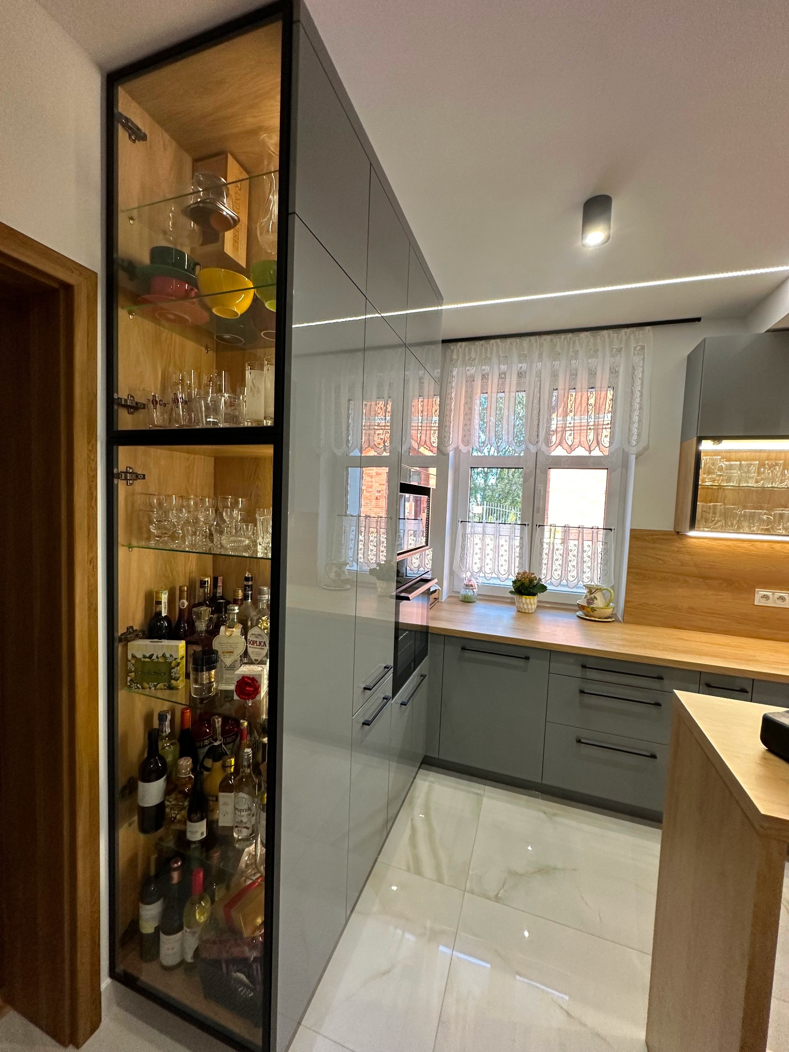 Szara kuchnia z wysoką, przeszkloną szafką wypełnioną szkłem i butelkami alkoholu, drewniany blat i białe płytki podłogowe.