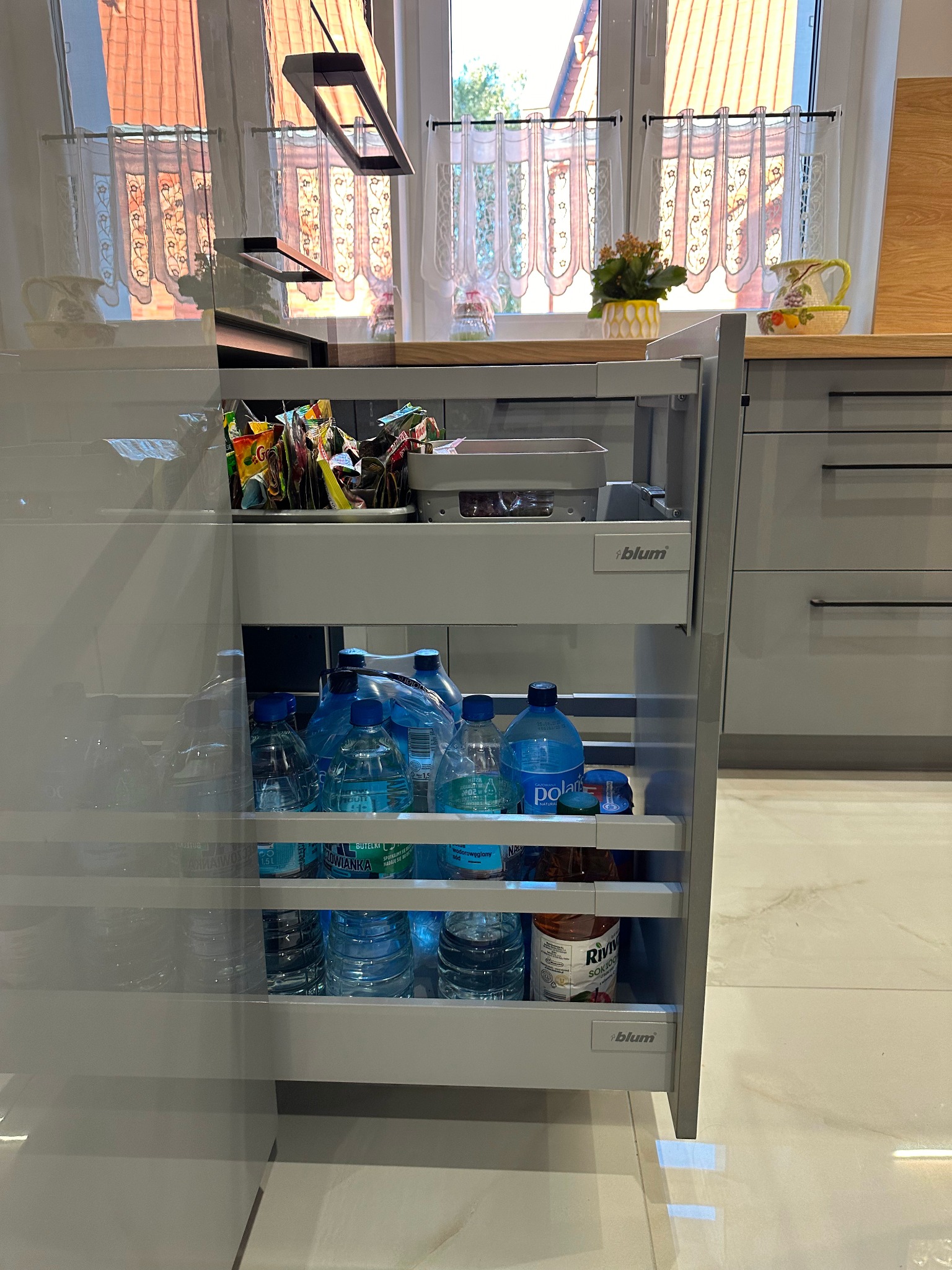 Wysuwana szafka kuchenna z szarymi frontami, wypełniona butelkami wody i przekąskami, z systemem Blum, widok na okno z dekoracyjnymi firankami i drewniany blat z ozdobami.
