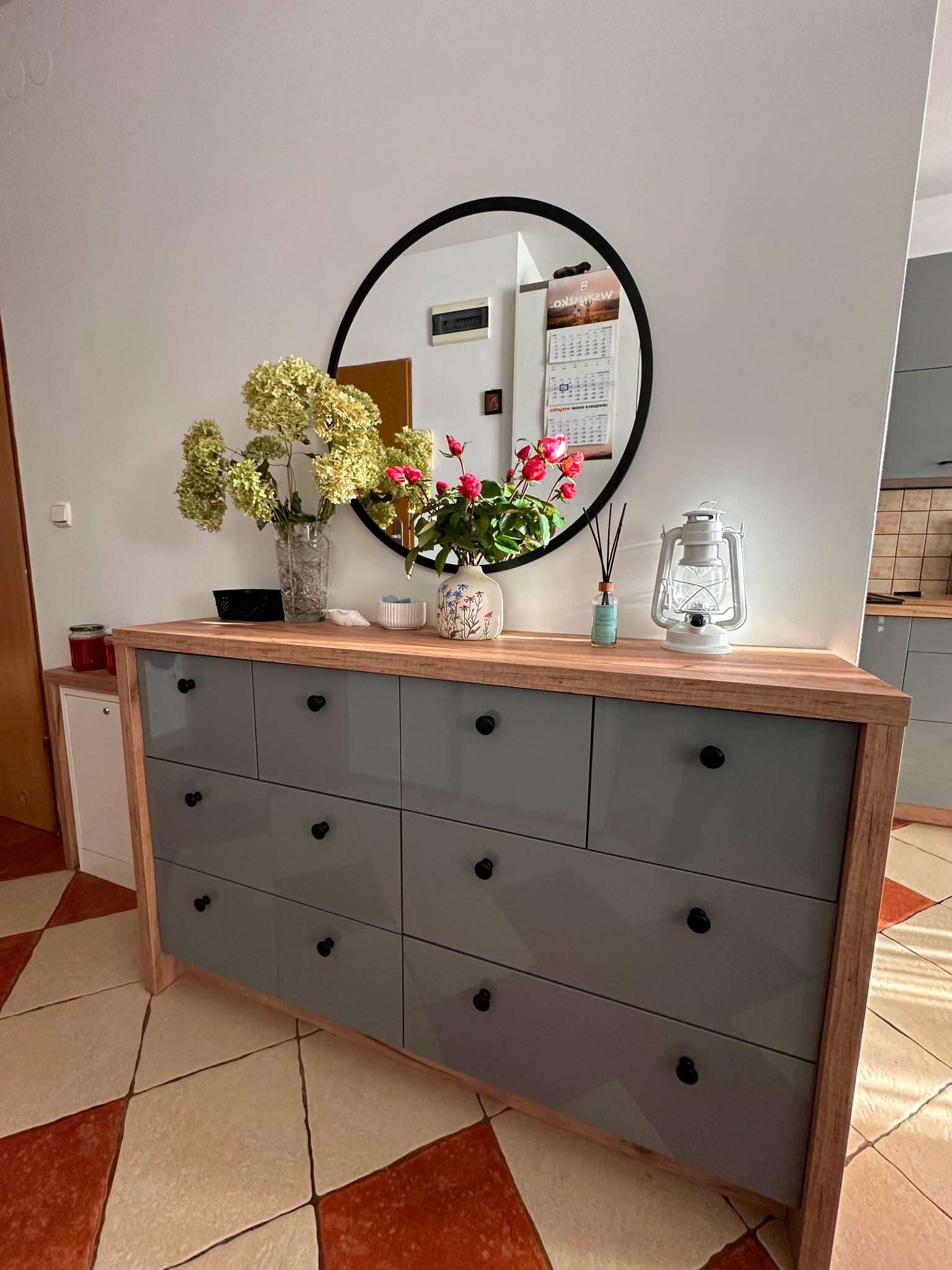 Szara komoda z drewnianym blatem i czarnymi uchwytami, na blacie wazony z kwiatami i lampion dekoracyjny, okrągłe lustro w tle, płytki podłogowe w romby.