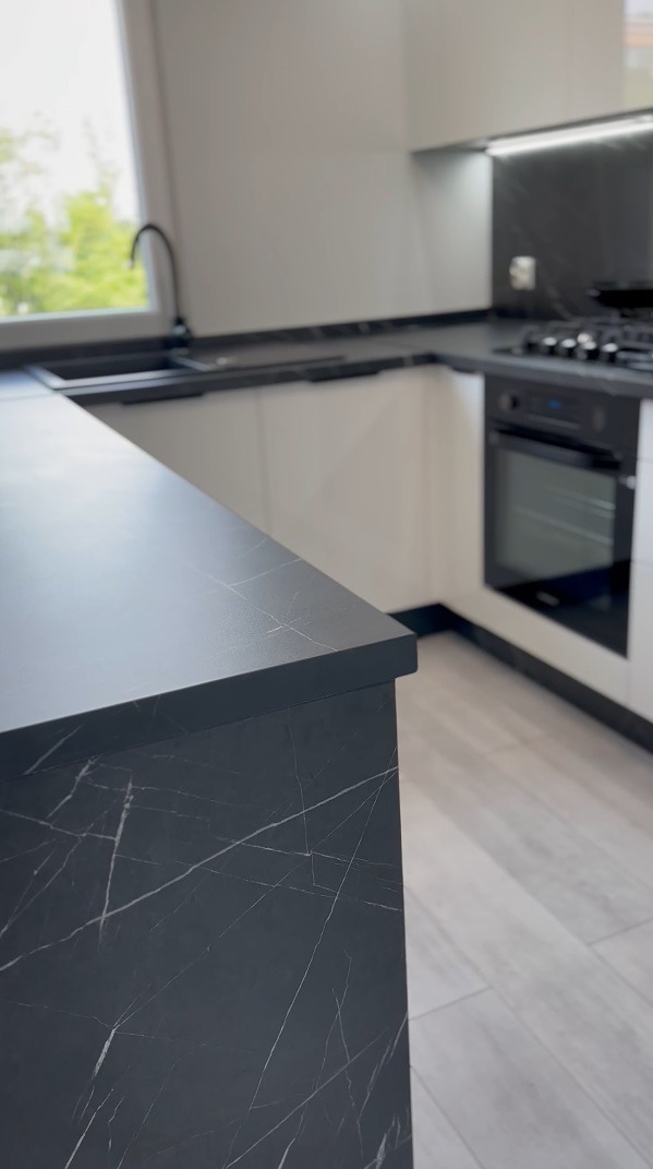 Czarny blat kuchenny z marmurowym wzorem, na tle białych szafek i sprzętów AGD w nowoczesnej kuchni.