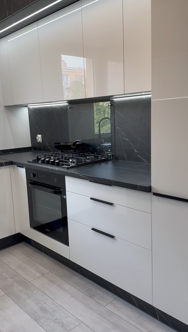 Nowoczesna kuchnia z białymi, lakierowanymi frontami, czarnym blatem z motywem marmuru i podświetleniem LED, widoczny piekarnik i płyta gazowa z patelnią.