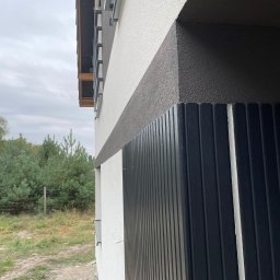 Renova Tomasz Januchowski - Fragment elewacji budynku z jasną i ciemną strukturą tynku oraz pionowymi panelami w kolorze ciemnego grafitu, widok z boku.