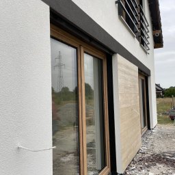 Renova Tomasz Januchowski - Elewacja budynku z oknami i dekoracyjnym panelem imitującym drewno. Widoczna struktura białej ściany, czarny pas dekoracyjny nad oknami. W tle słup wysokiego napięcia.