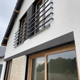 Renova Tomasz Januchowski - Elewacja budynku z oknami w drewnianych ramach, zabezpieczonymi czarnymi, metalowymi balustradami. Fragment ściany wykończony panelami imitującymi drewno, sąsiadujący z białą, chropowatą strukturą.