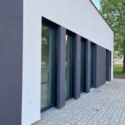 Renova Tomasz Januchowski - Elewacja budynku z szarą i białą gładzią, czarne ramy okienne, kostka brukowa przed budynkiem.