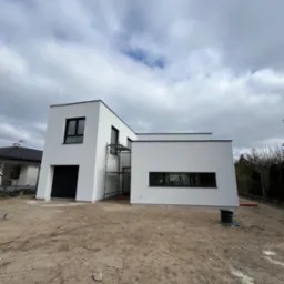 Nowoczesny, biały dom z płaskim dachem w trakcie budowy, widoczne rusztowania i niezagospodarowany teren wokół, pochmurne niebo w tle.