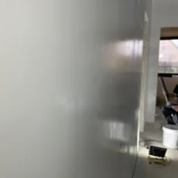 Świeżo położona, jasnoszara powierzchnia ściany z widocznym połyskiem, sugerującym mokrą jeszcze warstwę, z boku widoczny fragment pokoju w trakcie prac wykończeniowych, w tle kubeł i sprzęt...