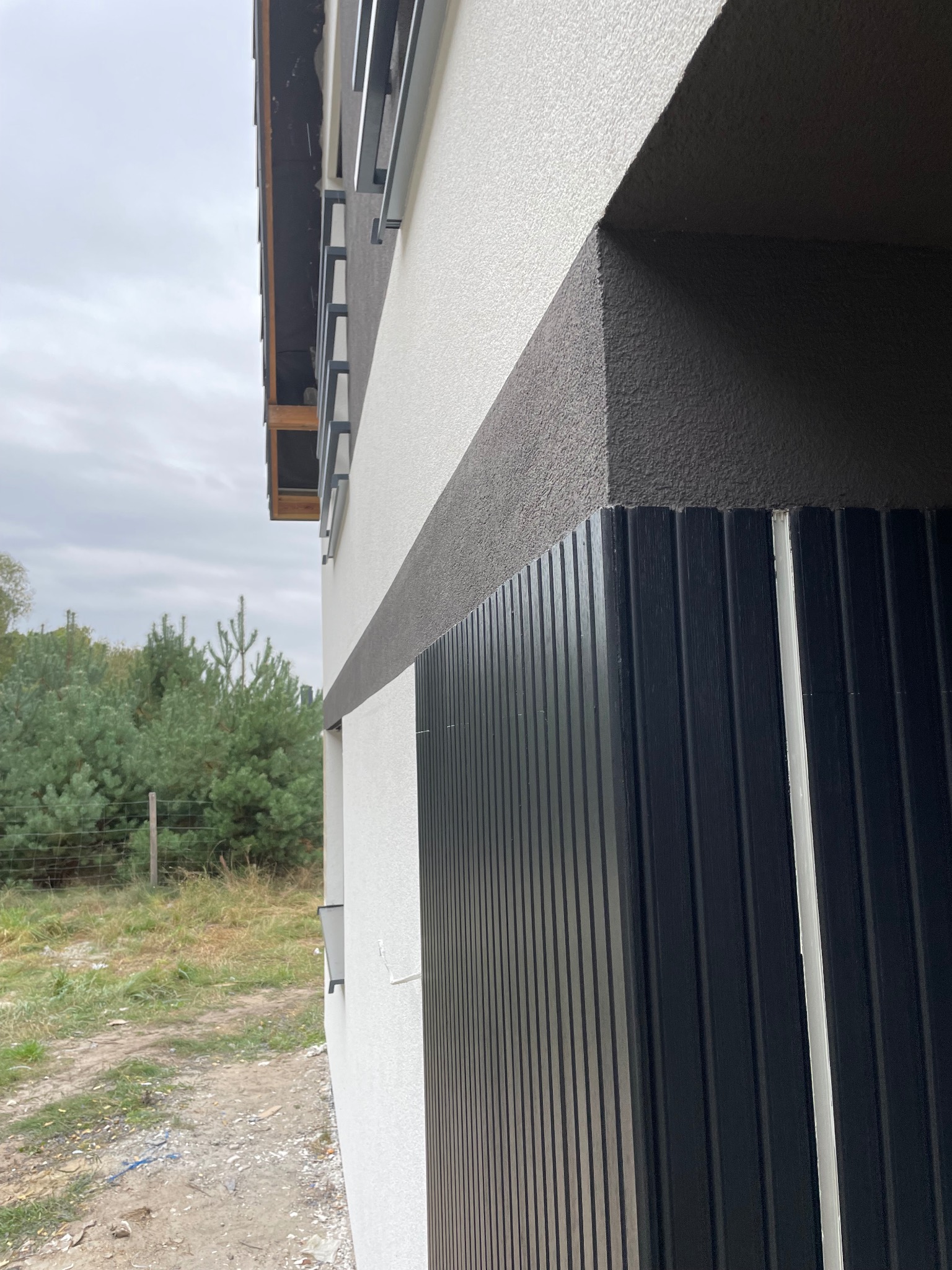 Fragment elewacji budynku z jasną i ciemną strukturą tynku oraz pionowymi panelami w kolorze ciemnego grafitu, widok z boku.