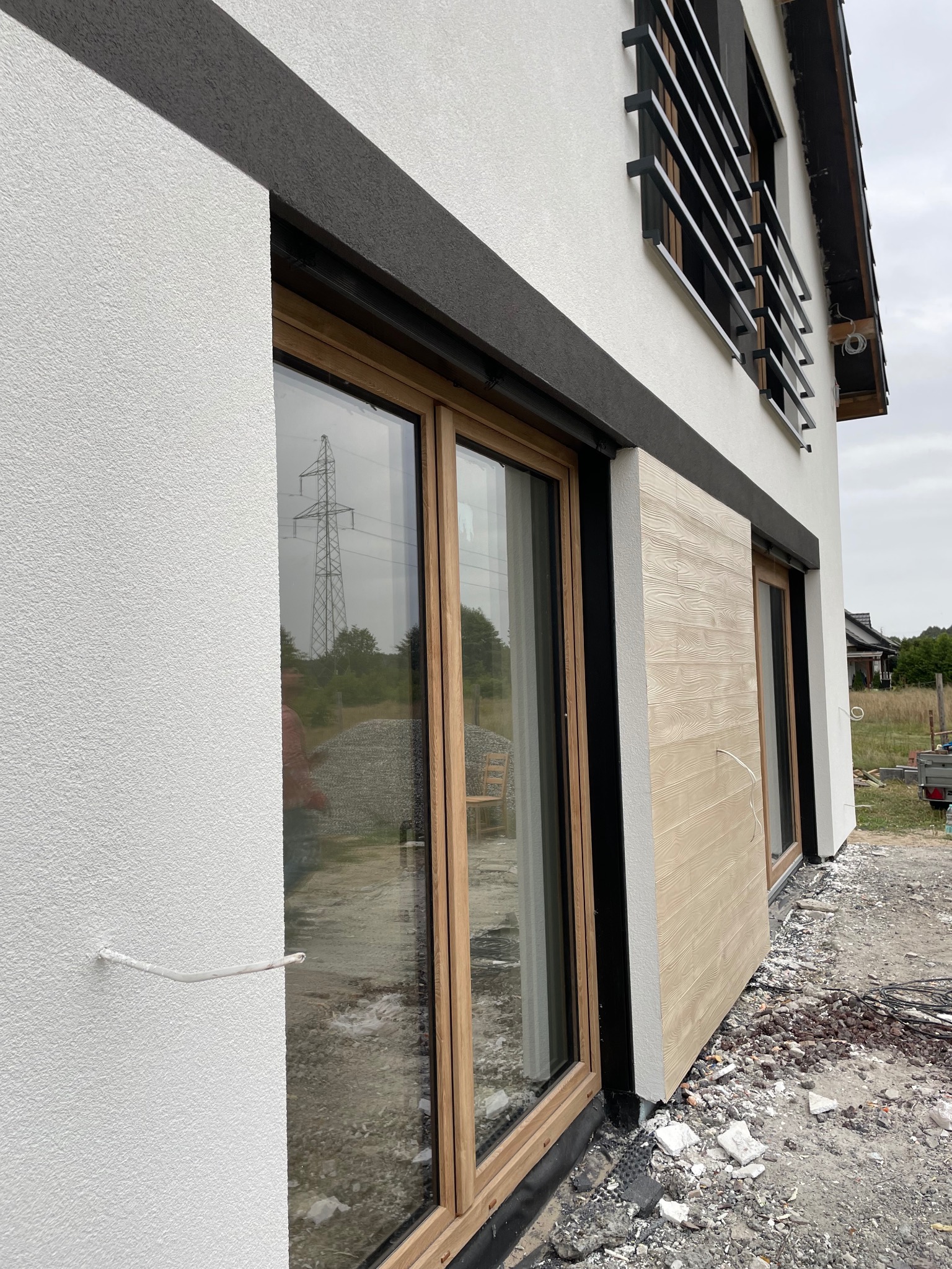 Elewacja budynku z oknami i dekoracyjnym panelem imitującym drewno. Widoczna struktura białej ściany, czarny pas dekoracyjny nad oknami. W tle słup wysokiego napięcia.