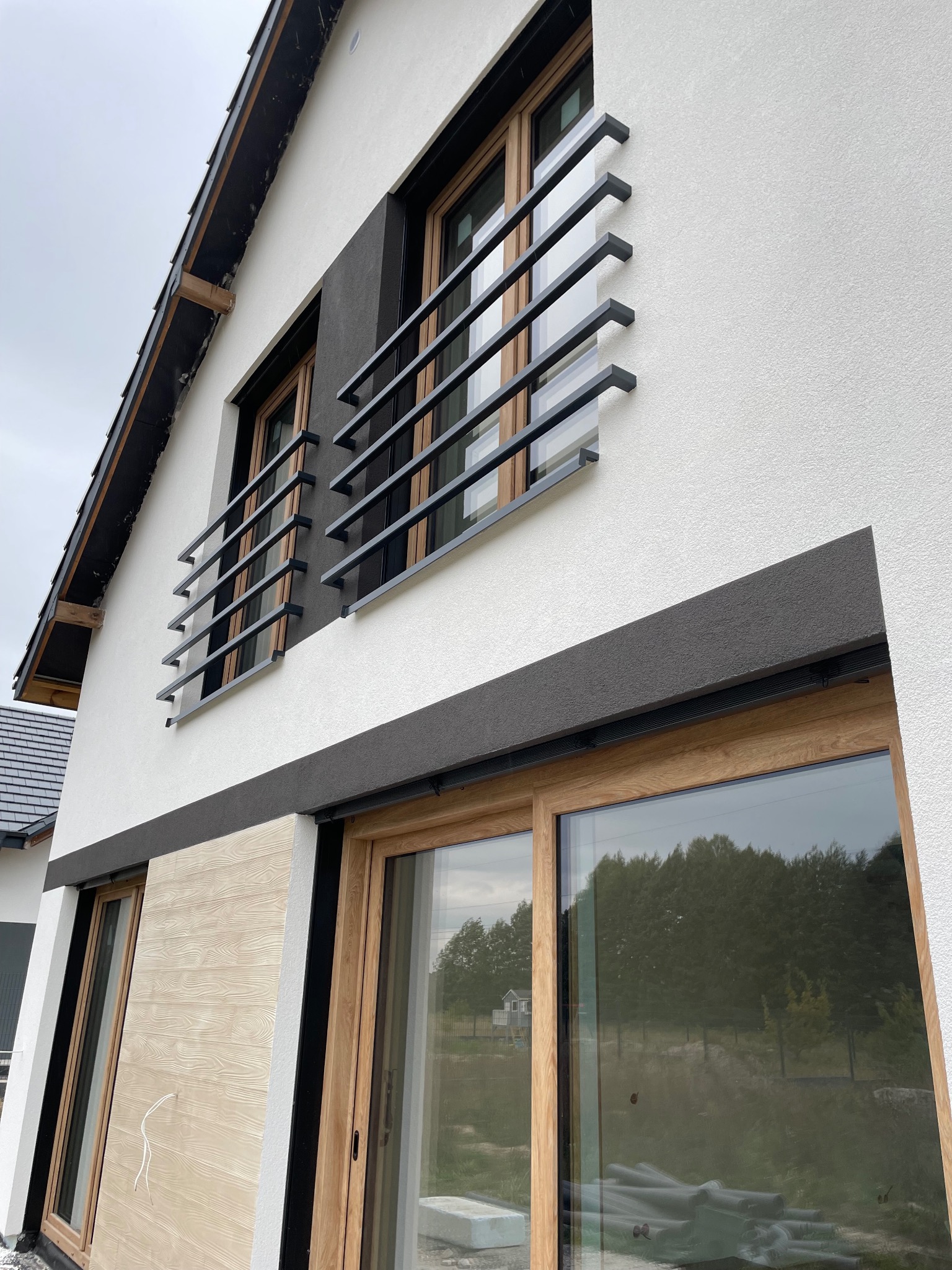 Elewacja budynku z oknami w drewnianych ramach, zabezpieczonymi czarnymi, metalowymi balustradami. Fragment ściany wykończony panelami imitującymi drewno, sąsiadujący z białą, chropowatą strukturą.