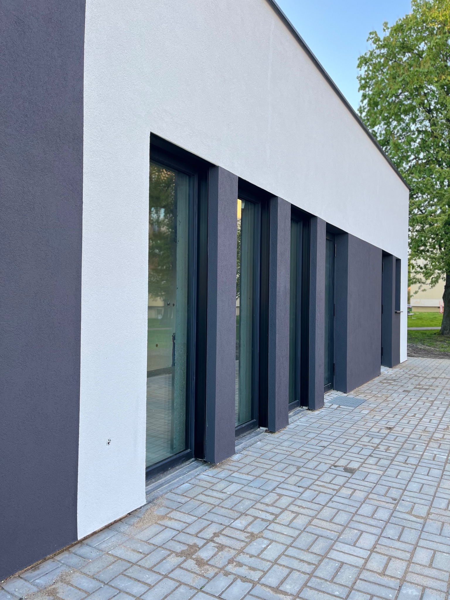 Elewacja budynku z szarą i białą gładzią, czarne ramy okienne, kostka brukowa przed budynkiem.