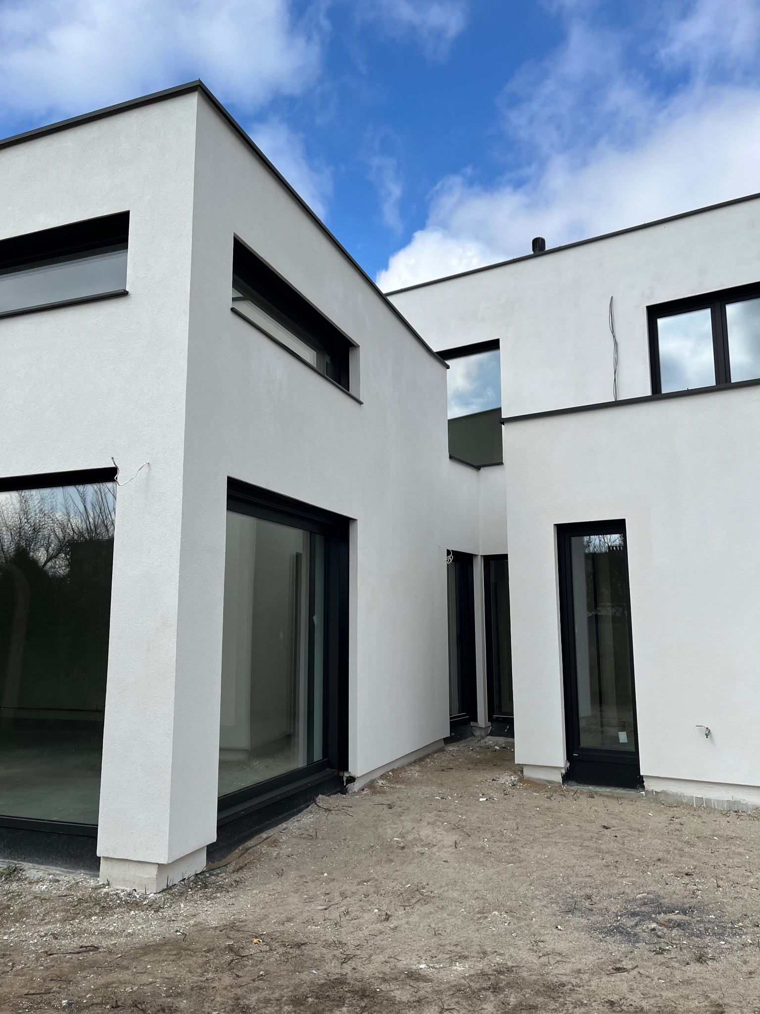 Nowoczesny, biały budynek z płaskim dachem i czarnymi ramami okiennymi, widok z zewnątrz na jeszcze niezagospodarowany teren.