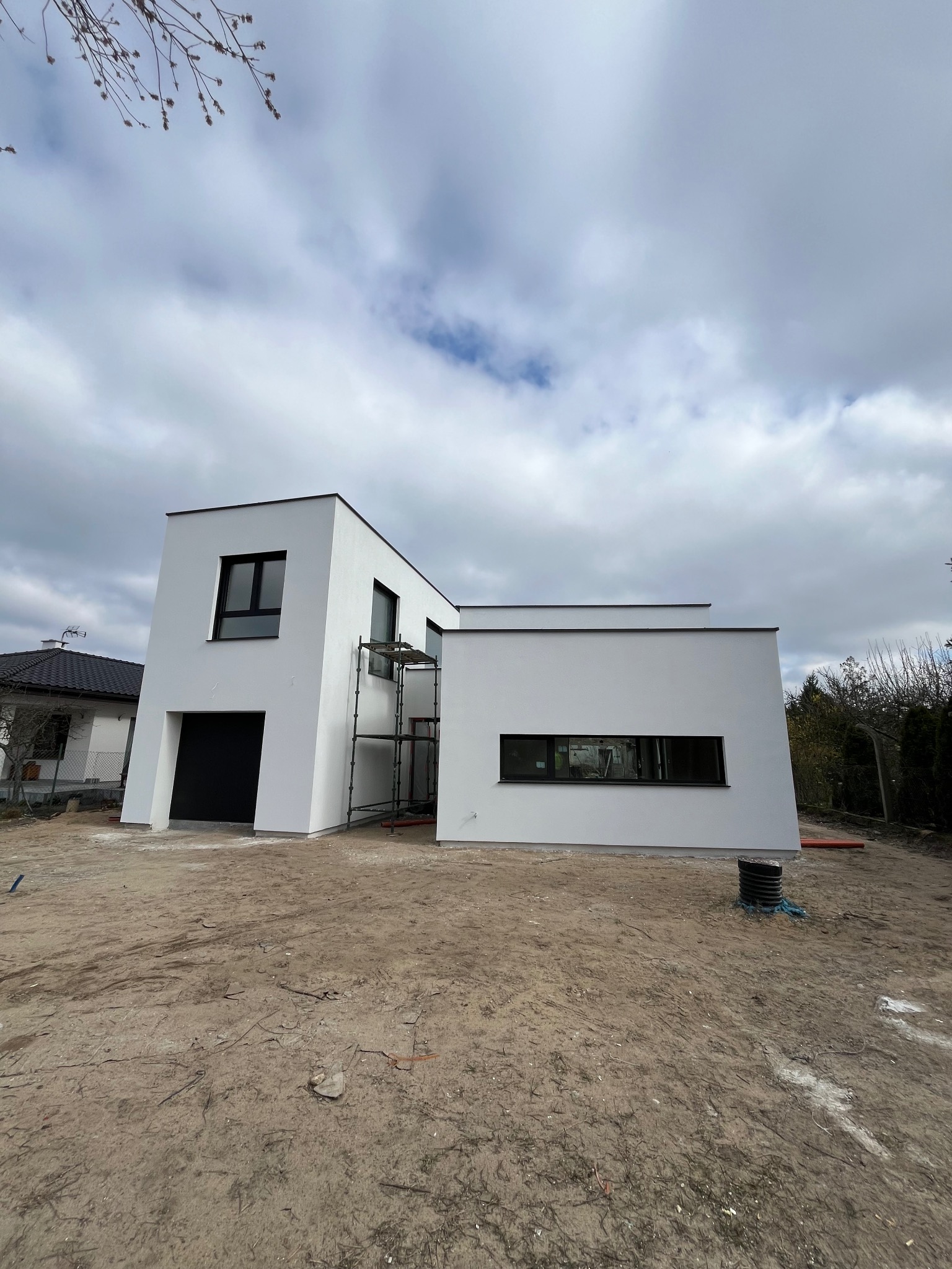 Nowoczesny, biały dom z płaskim dachem w trakcie budowy, widoczne rusztowania i niezagospodarowany teren wokół, pochmurne niebo w tle.