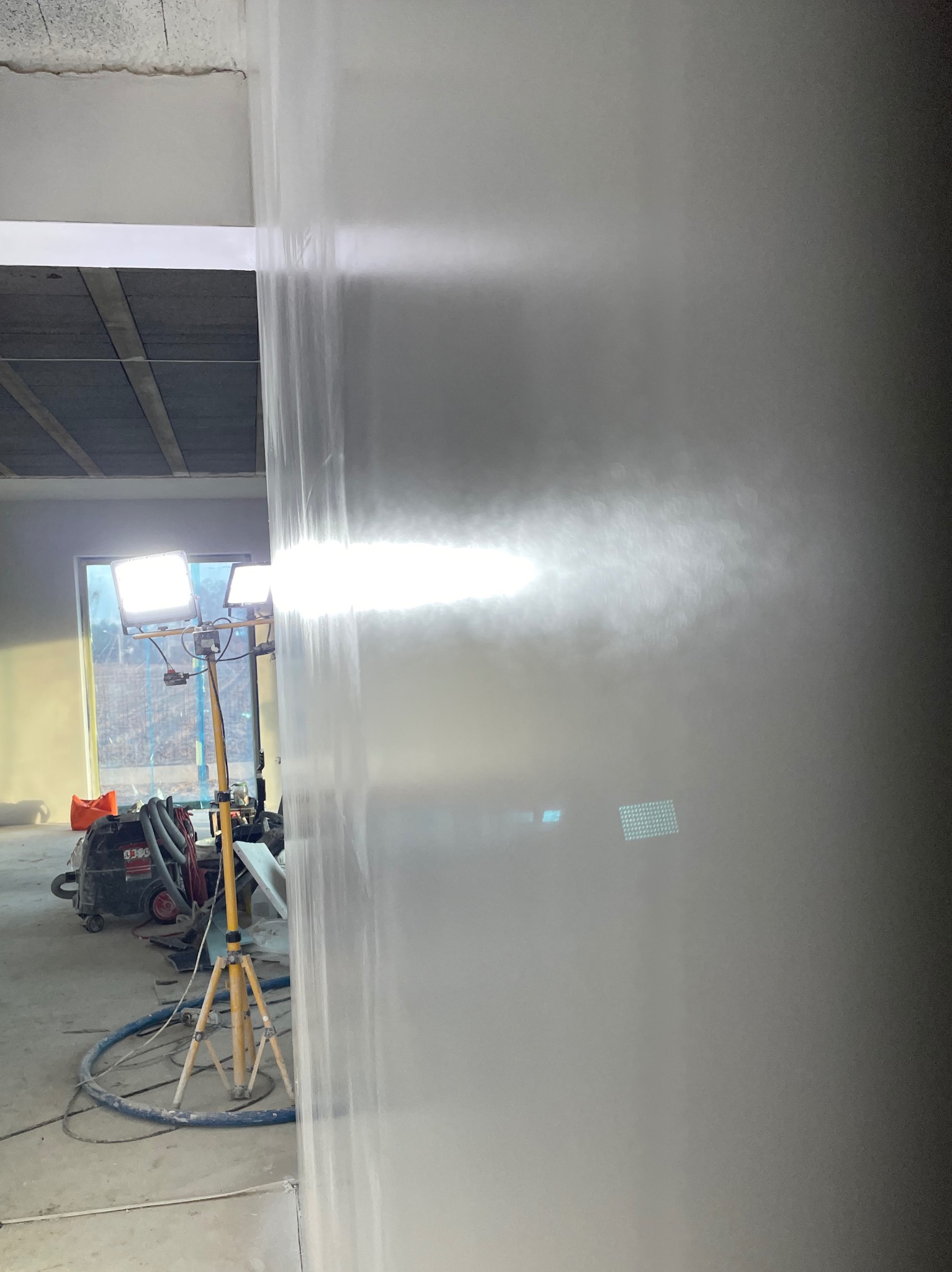 Świeżo nałożona, mokra gładź na ścianie w trakcie schnięcia, oświetlona lampami budowlanymi na statywie, z widocznym agregatem szpachlarskim i wężem na podłodze w surowym wnętrzu.