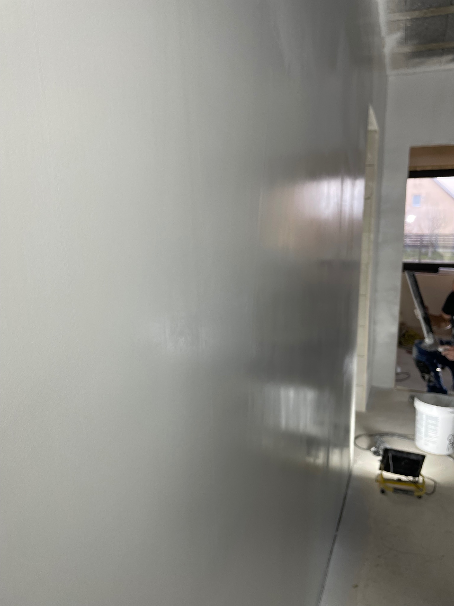 Świeżo położona, jasnoszara powierzchnia ściany z widocznym połyskiem, sugerującym mokrą jeszcze warstwę, z boku widoczny fragment pokoju w trakcie prac wykończeniowych, w tle kubeł i sprzęt...