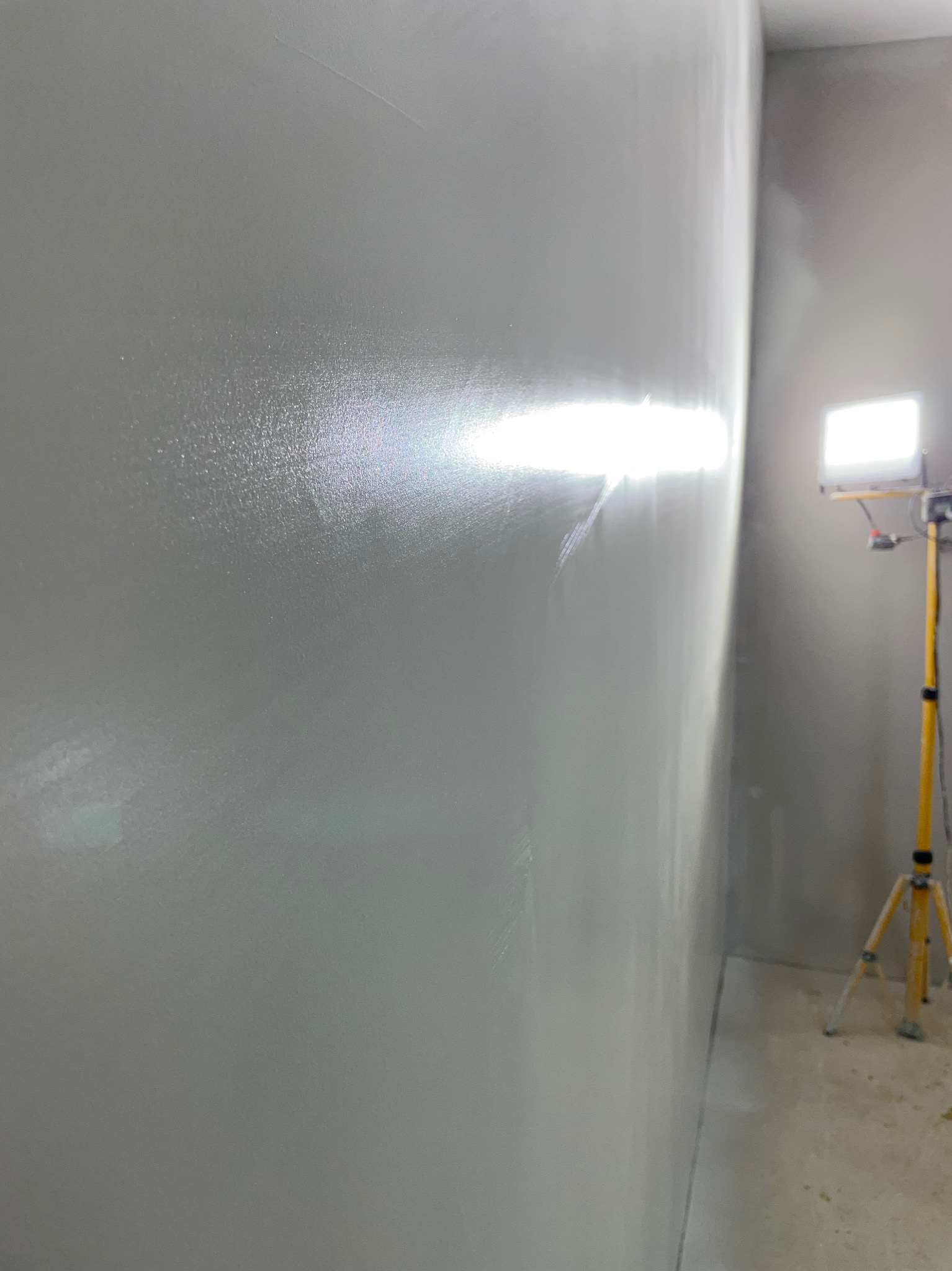 Świeżo położona, jasnoszara ściana w rogu pomieszczenia, oświetlona lampą budowlaną na żółtym statywie. Widoczne lekkie niedoskonałości powierzchni.