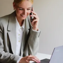Uśmiechnięta blondynka w szarym żakiecie rozmawia przez telefon i pracuje na laptopie, na biurku leży pomarańczowy notes.