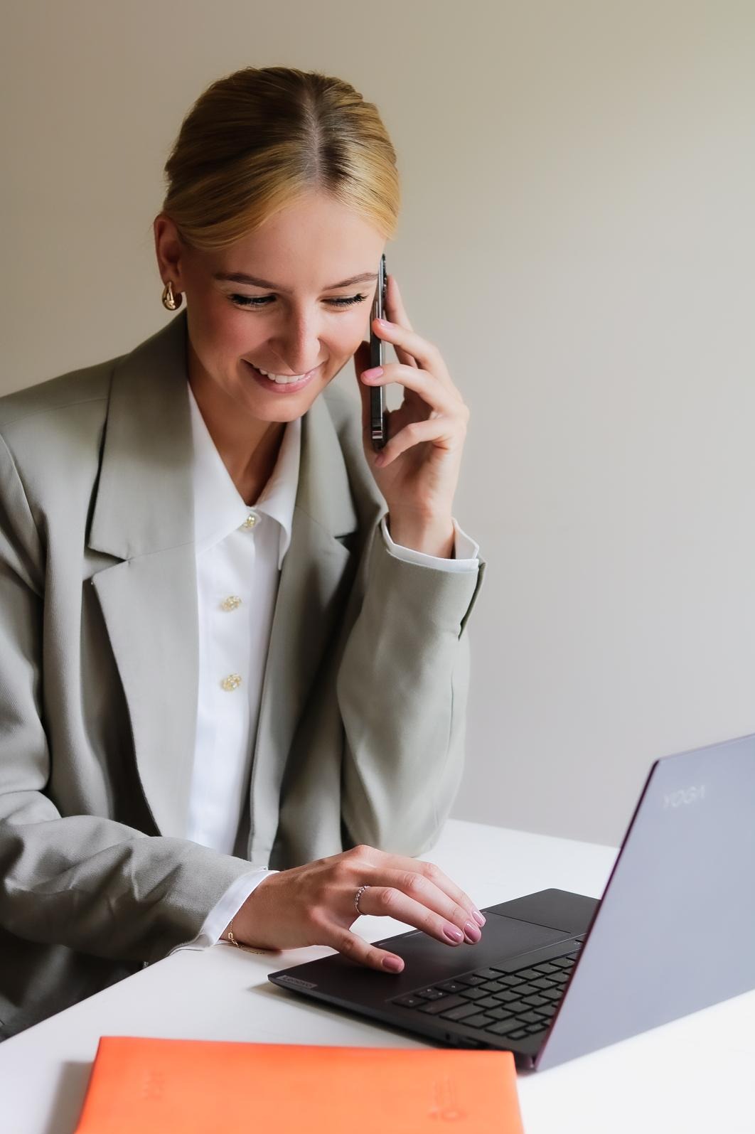 Uśmiechnięta blondynka w szarym żakiecie rozmawia przez telefon i pracuje na laptopie, na biurku leży pomarańczowy notes.