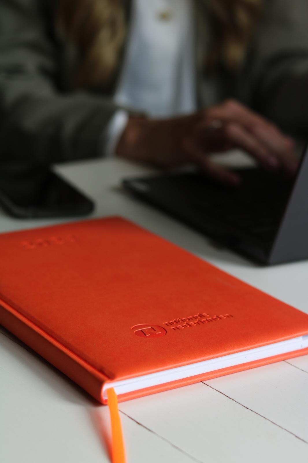 Pomarańczowy notatnik z wytłoczonym logo firmy ubezpieczeniowej na białym biurku, w tle osoba pracująca na laptopie.