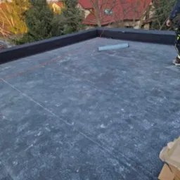 Płaski dach pokryty czarną papą, widoczne ślady stóp i rozłożona linka wyznaczająca linię, w tle drzewa i dom z czerwoną dachówką.