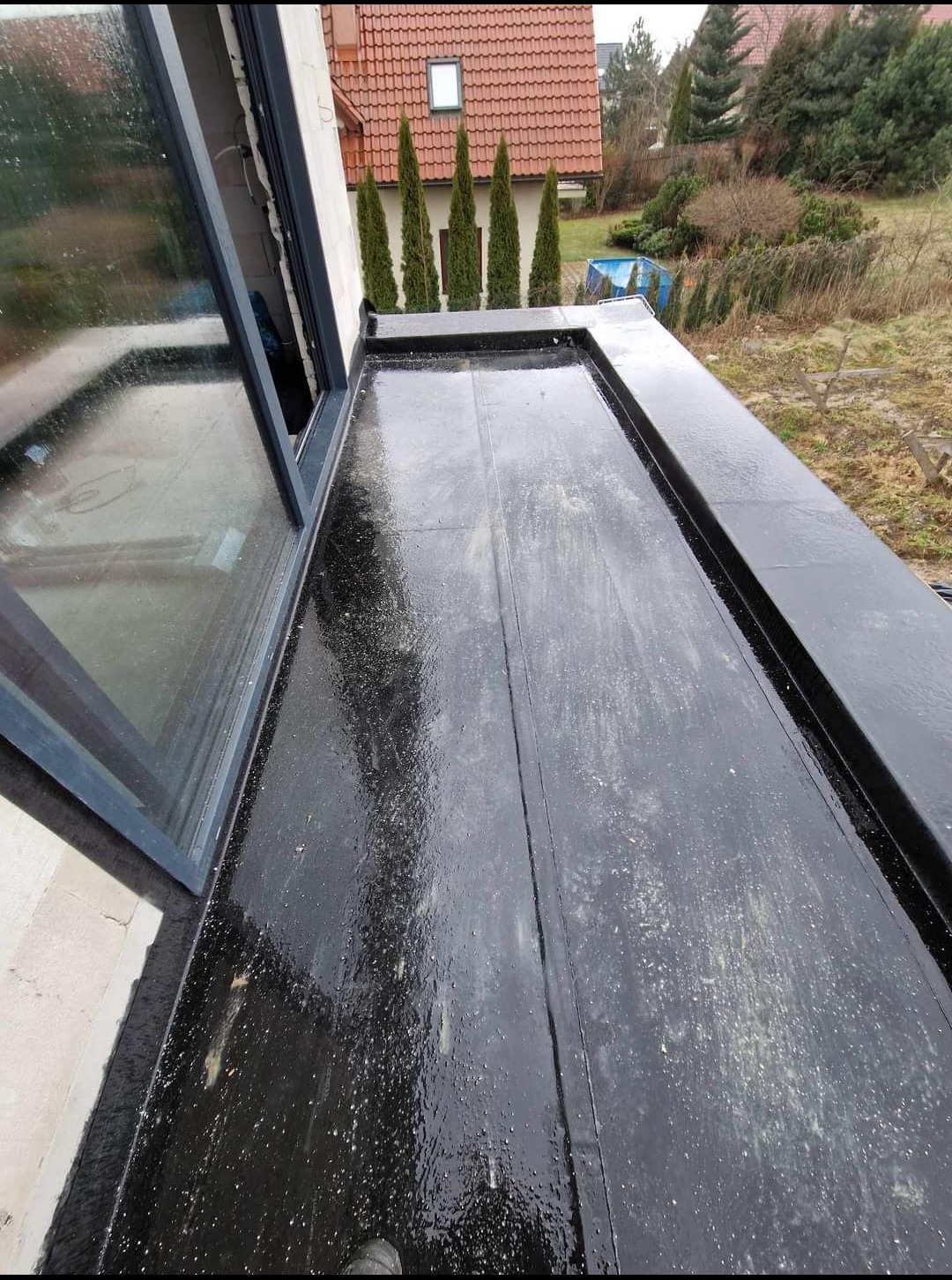 Czarny, płaski dach pokryty papą bitumiczną, widoczne łączenia i okno dachowe, na zewnątrz budynek mieszkalny i drzewa.
